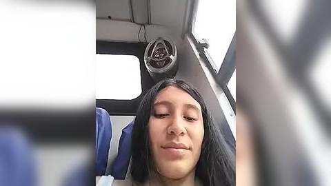 Media: Video of a serene young woman with long black hair, eyes closed, sitting in a bus. Background shows a window, a speaker, and a blue seat.