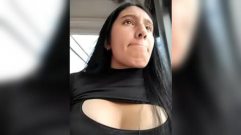 Media: Video of a woman with long black hair, wearing a tight black top with a large cutout revealing her ample cleavage, captured from a low angle, indoors.