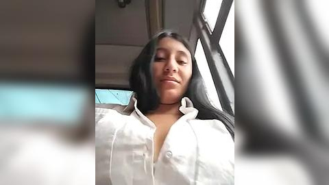 Media: Video of a woman with medium-length black hair and light skin, wearing a white, unbuttoned blouse, sitting in a car. The background features car seats and a blurred figure.