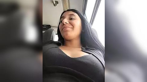 Media: Video of a young, light-skinned Black woman with long, straight black hair, smiling while seated in a car, wearing a black ribbed tank top.