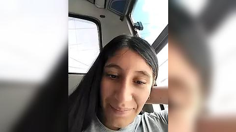 Media: Video of a young woman with long, straight black hair and fair skin, smiling while seated in a vehicle, wearing a light gray shirt. The background shows a blurred window and a partially visible person.