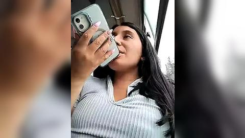 Media: Video of a young woman with long black hair, medium complexion, and medium build, wearing a gray ribbed knit top, taking a selfie in a car, blurred background.