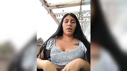 Media: Video of a young Latina woman with long black hair, wearing a light blue button-up blouse, sitting in a car. Background shows a white car and an out-of-focus person.