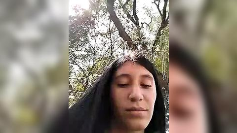 Media: Video of a young woman with long black hair, wearing a dark top, standing in a sunlit forest with dense green foliage and sunlight filtering through the trees.