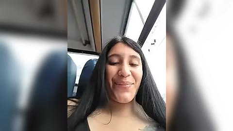 Media: Video of a young woman with long black hair, smiling with closed eyes, sitting in a bus with dark blue seats.