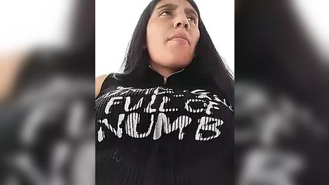 Media: Video of a young, fair-skinned woman with long black hair, wearing a black top with white, bold, handwritten text reading \"F*ck the Police,\" standing against a blurred background.