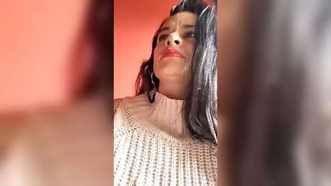 Media: Video of a young woman with long black hair, wearing a beige knitted sweater, captured from a low angle, against a blurred red background.