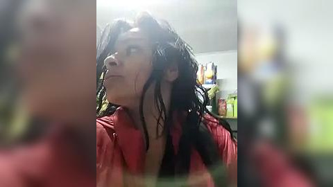 Media: A blurred video of a person with wet, dark, tangled hair, wearing a red shirt, standing in a dimly lit room with shelves in the background.