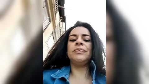 Media: Video of a woman with long black hair, wearing a blue denim shirt, standing in front of a brown building with closed shutters, looking upwards with a serene expression.