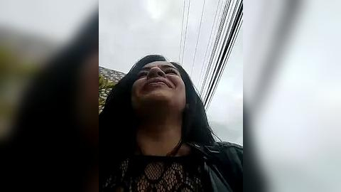 Media: A video of a smiling woman with long black hair and a black lace top, taken from a low angle, with power lines and a cloudy sky in the background.