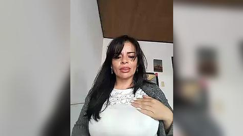 Media: Video of a woman with long black hair, medium skin tone, wearing a white lace top and grey cardigan, standing in a modern kitchen with white cabinets and wooden ceiling.