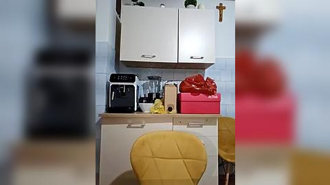 Media: A video of a modern kitchen with white cabinets, a yellow chair, a black coffee maker, a pink gift box, and a tiled backsplash.