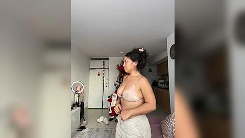 Media: Video of a woman with medium brown skin, dark hair in a bun, wearing a sheer white bra and high-waisted pants, standing in a modern, sparsely furnished living room with a white door and a red couch.