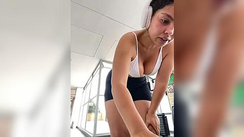 Media: A video of a fit, dark-haired woman with large breasts in a white tank top and black shorts, exercising on a treadmill in a modern gym.