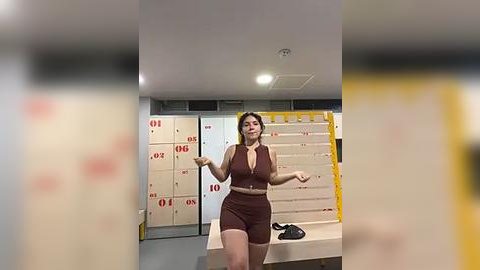 Media: Video of a plus-size woman with dark hair in a maroon sports bra and shorts, gesturing in a locker room with beige lockers labeled \"01\" and \"06,\" and a yellow storage rack in the background.