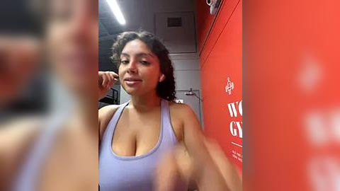 Media: Video of a young woman with curly hair, medium skin tone, wearing a light blue sports bra, standing in a gym with red walls and white text.