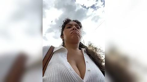 Media: Video of a woman with curly hair, wearing a low-cut white blouse, looking up at the sky with a pensive expression.