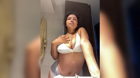 Media: Video of a curvy, light-skinned woman with curly hair in a white bra and panties, covering her chest in a bathroom with beige tiles, a towel, and a dark wooden door.