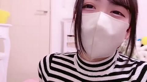 Media: Video of an Asian woman with straight black hair, wearing a white face mask and black-and-white striped sweater, standing in a modern, white-walled room with laundry baskets and a yellow towel visible in the background.