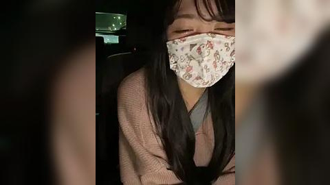 Media: Video of an Asian woman with long black hair, wearing a floral mask and a beige sweater, seated in a dark car.