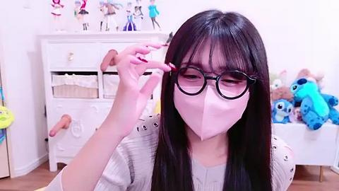 Media: A video of an Asian woman with long black hair, wearing glasses and a pink mask, applying eyelashes in a bedroom with stuffed toys and a white dresser.