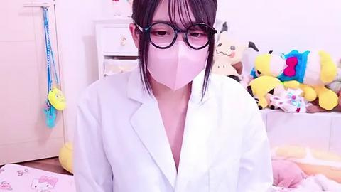 Media: A video of an Asian woman with long black hair, wearing large glasses and a white lab coat, seated in a room filled with plush toys, including a large, yellow plush bear.