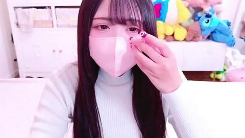 Media: Video of an Asian woman with long black hair, wearing a light blue sweater and a pink face mask, holding a pink phone, in a brightly lit, plush-filled room with toys and a white bed.