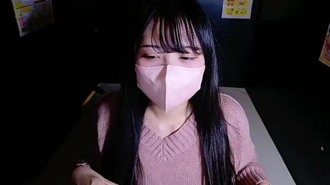 Media: Video of an East Asian woman with long black hair and pale skin, wearing a pink face mask and a pink sweater, sitting at a desk in a dimly lit room.