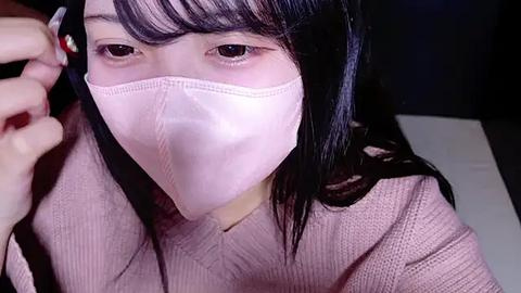 Media: Video of an Asian woman with long black hair, wearing a light pink face mask and a matching sweater, adjusting her hair with one hand. The background is dark, with a hint of a white surface.