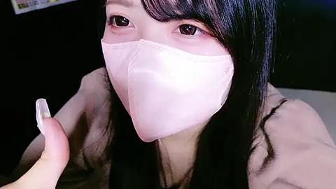 Media: Video of an Asian woman with long black hair, wearing a white face mask, holding a white nail file. She has light skin, brown eyes, and is indoors with a blurred background.