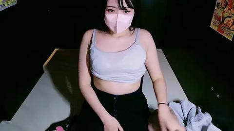 Media: Video of a young Asian woman with fair skin, wearing a white mask, light blue crop top, and black skirt, sitting at a table. A hand reaches towards her.