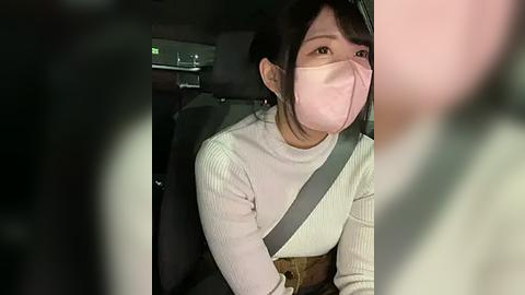 Media: Video of an East Asian woman wearing a face mask, gray shoulder strap, white sweater, and brown belt, sitting in a dark car interior with blurred figures in the foreground.