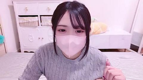 Media: Video of an Asian woman with short black hair, wearing a light gray ribbed sweater and a pink face mask, standing in a white bedroom with a bed and white dresser.