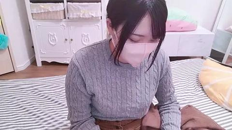 Media: Video of an Asian woman with straight black hair, wearing a gray knitted sweater, face mask, and headphones, sitting on a striped bed in a pastel-colored room.