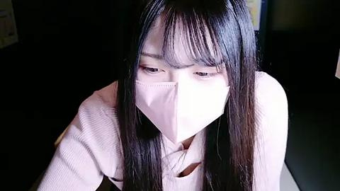 Media: Video of a young Asian woman with long black hair, wearing a white face mask, looking down. She has light skin and is dressed in a beige sweater. Background is dark and blurred, suggesting an indoor setting.