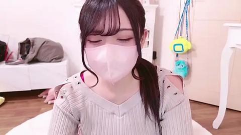 Media: Video of a young Asian woman with long black hair, wearing a light gray ribbed sweater and a white surgical mask, sitting on a white rug in a cluttered room with a white dresser and cat toys.