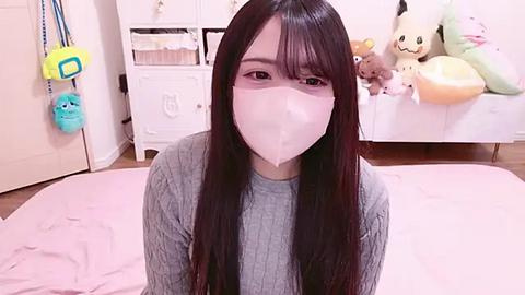 Media: Video of an Asian woman with long black hair, wearing a white face mask, gray sweater, and pink blanket in a pastel-colored bedroom with stuffed toys and a white dresser.