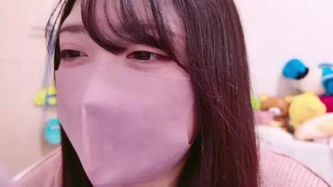 Media: Video of an Asian woman with long, straight black hair and fair skin wearing a light pink, ribbed long-sleeved shirt, with a light pink surgical mask covering her nose and mouth, slightly obscuring her eyes. The background is blurry, showing a cluttered room with colorful toys.