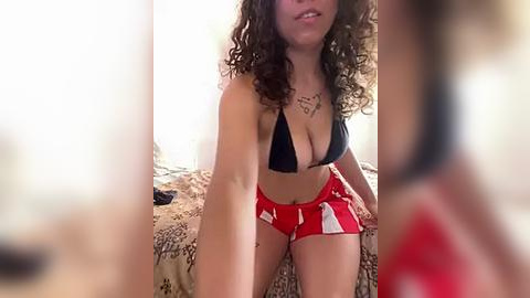 Media: Video of a young, light-skinned woman with curly hair, wearing a black bikini top and red shorts, sitting on a bed with a patterned sheet in a dimly lit room.