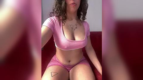 Media: Video of a light-skinned woman with curly brown hair, wearing a pink crop top and shorts, sitting on a red couch.