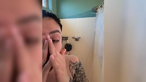 Media: Video of an Asian man with short black hair, wearing a grey shirt, covering his face with his hands, standing in a shower with white tiles and a green wall, partially obscured by a white curtain.