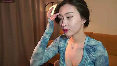 Media: A video of an East Asian woman with short black hair, wearing a low-cut, blue tie-dye top, adjusting her hair in a cozy, dimly-lit room.