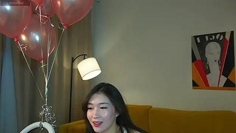 Media: Video of an Asian woman with long black hair, fair skin, and a red dress, holding a cluster of red balloons, seated in a modern living room with a mustard-yellow couch, a black floor lamp, and a colorful abstract painting on the wall.