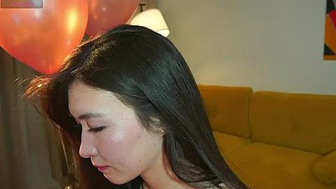 Media: Video of a young woman with long dark hair, pale skin, and pink blush, holding a red balloon, standing next to a mustard-yellow couch in a warmly lit room.