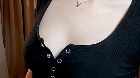 Media: Video of a woman's upper chest, showing a black ribbed top with a deep V-neckline and visible cleavage, revealing fair skin. The background features brown curtains.