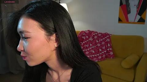 Media: Video of an Asian woman with long black hair, wearing a black top, in a modern living room with a mustard-yellow couch, red floral pillow, and abstract artwork.