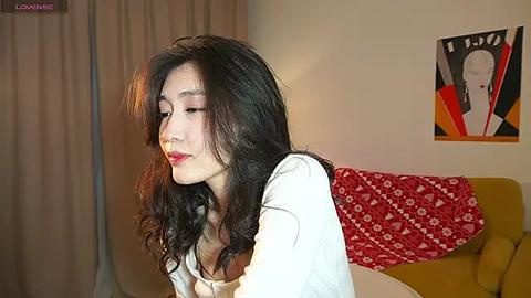 Media: Video of an Asian woman with long, wavy black hair, wearing a white top, leaning against a mustard-yellow couch with a red floral blanket. She has a serene expression.