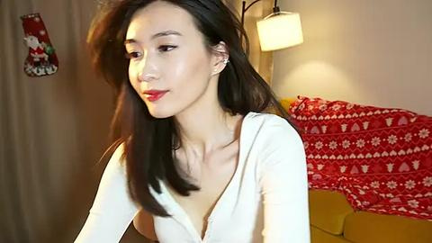 Media: Video of an East Asian woman with long black hair, fair skin, wearing a white, deep V-neck top, red lipstick, standing indoors, festive red and white patterned blanket behind her.