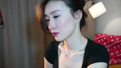 Media: Video of a young East Asian woman with fair skin, dark hair, and red lipstick, wearing a black top, sitting indoors with a lamp and red-patterned blanket in the background.