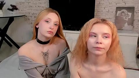 Media: Video of two young, fair-skinned, blonde women with tattoos, one wearing a black choker and gray hoodie, the other topless, sitting on a bed in a minimalist, modern room.